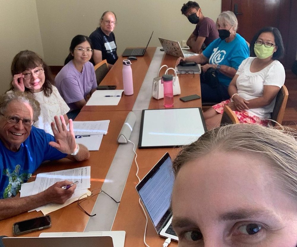 selfie of eight people, half of whom are wearing face masks, sitting around a long conference table with laptops, notes, and water bottles