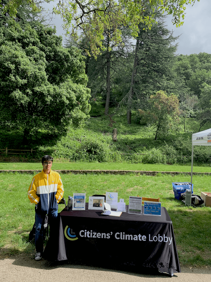 Photo of a young man standing next to a table with a tablecloth that says 'Citizens Climate Lobby' with green grass and trees in the background