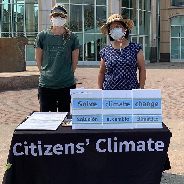 Photo of two women wearing face coverings standing behind a table that has a sign that says 'Solve Climate Change' and a tablecloth that says 'Citizens Climate'